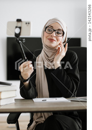 A smiling Muslim woman wearing a hijab and glasses is recording a video on her smartphone with a selfie stick while sitting at her desk 142543266