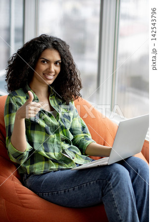 A young woman with curly hair sits comfortably in a beanbag chair, holding a laptop and giving a thumbs-up gesture with a smile A young woman with curly hair sits comfortably in a beanbag chair, holding a laptop and giving a thumbs-up gesture with a smile 142543796