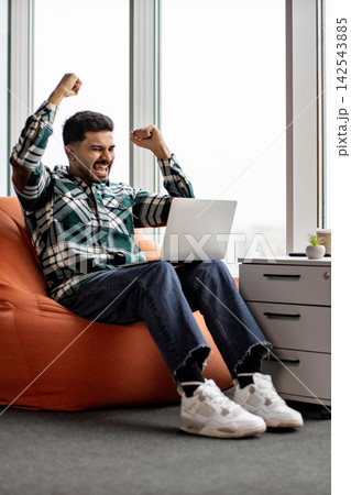 A young man in casual attire expresses joy and triumph while working on his laptop, seated comfortably in a beanbag chair A young man in casual attire expresses joy and triumph while working on his laptop, seated comfortably in a beanbag chair 142543885
