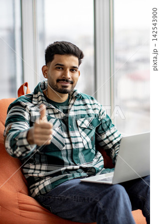 A young man with a beard and dark hair smiles while giving a thumbs-up gesture with a laptop on his lap 142543900