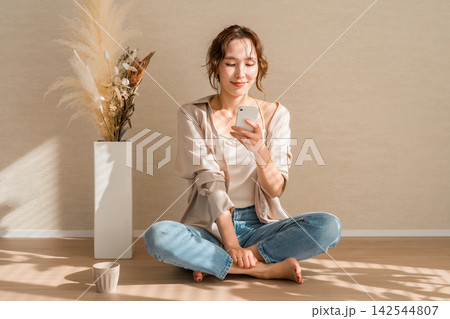 自然光が差し込む部屋でスマホを見ながらくつろぐ女性 自然光が差し込む部屋でスマホを見ながらくつろぐ女性 142544807