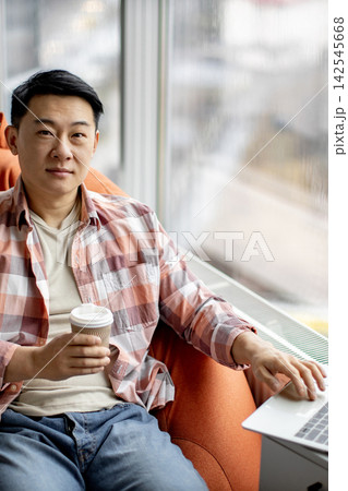 A man sits comfortably in an armchair by the window, holding a coffee cup and working on his laptop A man sits comfortably in an armchair by the window, holding a coffee cup and working on his laptop 142545668