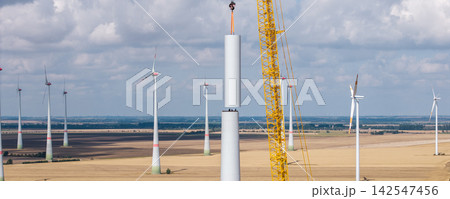 Aerial drone view wind turbine installation across harvested farmland, with cranes lifting tower sections. Renewable energy construction expands wind farm infrastructure under dramatic summer clouds Aerial drone view wind turbine installation across harvested farmland, with cranes lifting tower sections. Renewable energy construction expands wind farm infrastructure under dramatic summer clouds 142547456