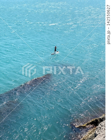 Lone paddleboarder crosses turquoise coastal water on bright sunny summer day. 142550627