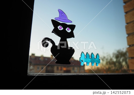 Silicone stickers for sticking to window glass or mirrors. Halloween theme. Black cat, purple witch hat, and a blue transparent fence. Room decor. In background is apartment building and evening sky Silicone stickers for sticking to window glass or mirrors. Halloween theme. Black cat, purple witch hat, and a blue transparent fence. Room decor. In background is apartment building and evening sky 142552073