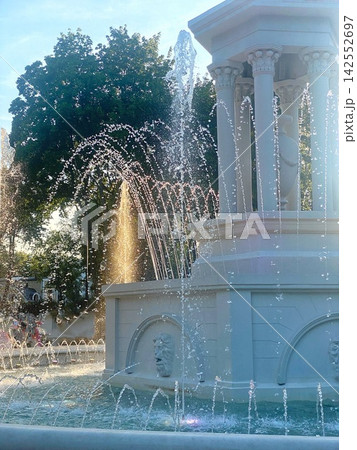 Elegant white fountain with sparkling water jets in a sunlit city park at golden hour. Elegant white fountain with sparkling water jets in a sunlit city park at golden hour. 142552697