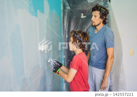 Father and son painting a wall together during home renovation indoors with paint rollers blue wall teamwork DIY repair family help and practical household improvement. Father and son painting a wall together during home renovation indoors with paint rollers blue wall teamwork DIY repair family help and practical household improvement. 142557221