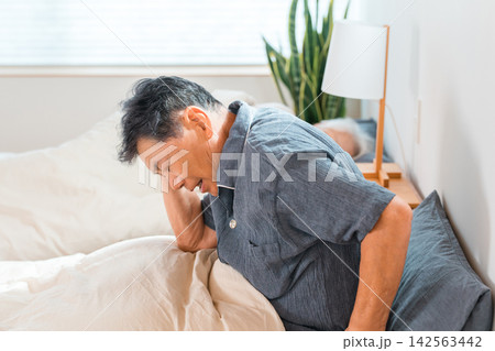 ベッドで頭を抱える60代から70代のシニア男性。寝起きの疲労や体調不良に悩むイメージ ベッドで頭を抱える60代から70代のシニア男性。寝起きの疲労や体調不良に悩むイメージ 142563442