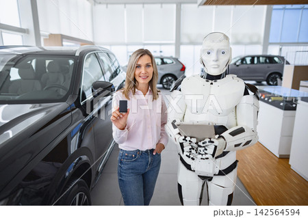 A woman holds a car key while standing next to a humanoid robot in a modern showroom with vehicles on display A woman holds a car key while standing next to a humanoid robot in a modern showroom with vehicles on display 142564594