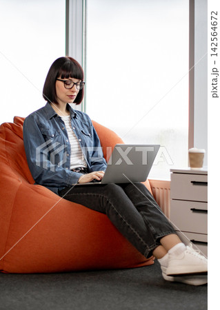 A young woman with glasses and a bob haircut sits comfortably on an orange bean bag chair, typing on her laptop with a coffee cup nearby A young woman with glasses and a bob haircut sits comfortably on an orange bean bag chair, typing on her laptop with a coffee cup nearby 142564672