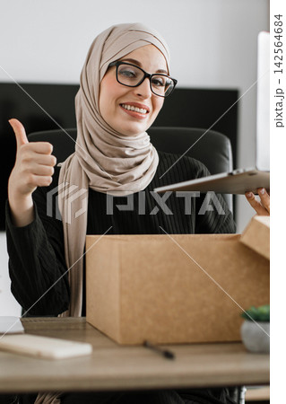 A smiling woman wearing a hijab and glasses gives a thumbs-up while holding a laptop and looking at an open cardboard box on her desk 142564684