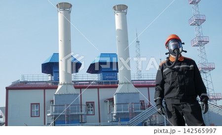 Protective suited man stands before industrial plant, wearing gas mask and helmet with focused expression. Power station towers and chimneys rise behind worker in harsh outdoor safety setting. Protective suited man stands before industrial plant, wearing gas mask and helmet with focused expression. Power station towers and chimneys rise behind worker in harsh outdoor safety setting. 142565997