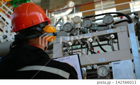Back view of maintenance worker in hard hat reviews gauge readings at industrial panel, focused and attentive. Worker stands before pipes, dials, and clipped paperwork, inspecting meters and valves. 142566011