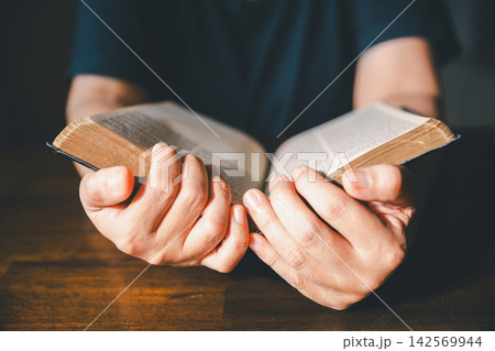 Close-up of hands turning pages of an old Bible on a wooden table, representing Christian scripture reading, faith study, devotion, worship, spiritual learning, reflection, wisdom, and religion. 142569944