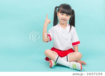 Smiling little girl in sports uniform sitting cross-legged and pointing finger up at copy space. 142571359