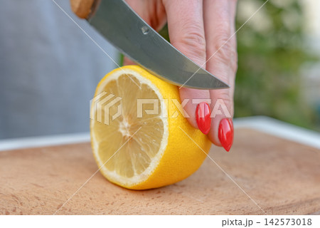 Woman's hand with red fingernails holding and slicing a fresh yellow lemon on a wooden cutting board, preparing it for cooking or juicing with a sharp knife 142573018
