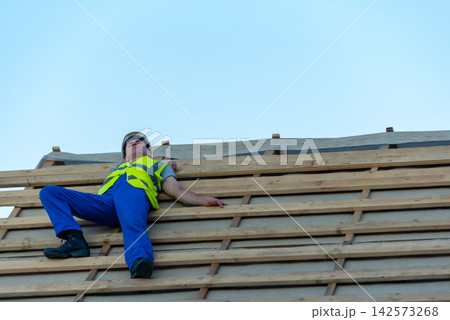 Construction worker in safety gear collapsed on a roof under construction, illustrating workplace accident, fall risk, injury potential and need for safety and prevention measures on job sites Construction worker in safety gear collapsed on a roof under construction, illustrating workplace accident, fall risk, injury potential and need for safety and prevention measures on job sites 142573268