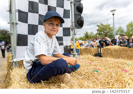 Boy watches soap box racing from hay bales beside checkered flag, enjoying outdoor community event in Germany. Young spectator rests near finish area while crowd gathers around downhill race course Boy watches soap box racing from hay bales beside checkered flag, enjoying outdoor community event in Germany. Young spectator rests near finish area while crowd gathers around downhill race course 142575557