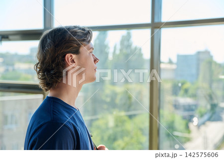 Pensive young man looking out large window with city view in profile Pensive young man looking out large window with city view in profile 142575606