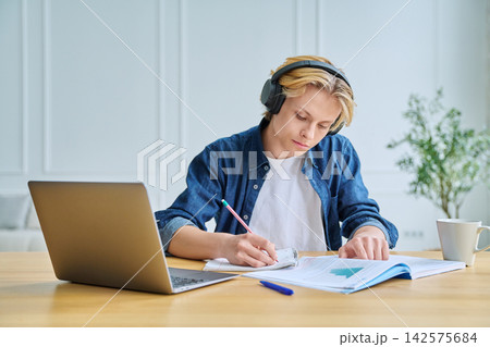 Teenage student in headphones writing at desk indoor Teenage student in headphones writing at desk indoor 142575684