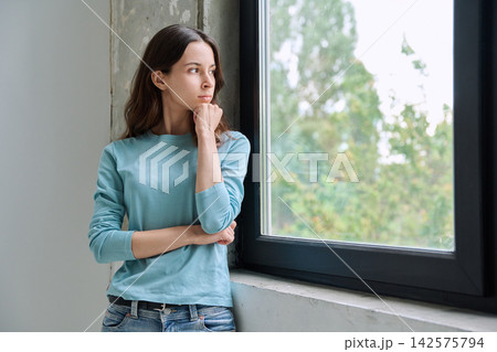 Thoughtful teen girl holding chin and looking out window 142575794