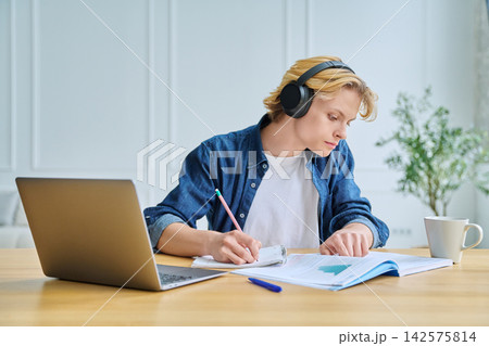 Teenage student in headphones writing at desk indoor Teenage student in headphones writing at desk indoor 142575814