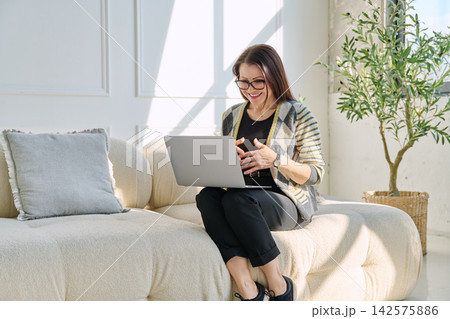 Smiling mature woman having video call on laptop while sitting on sofa in bright living room Smiling mature woman having video call on laptop while sitting on sofa in bright living room 142575886