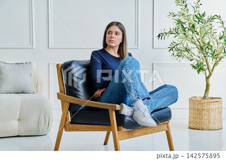 Portrait of thoughtful, pensive young woman sitting in chair at home Portrait of thoughtful, pensive young woman sitting in chair at home 142575895