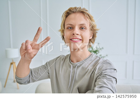 Happy teenage boy smiling at camera and showing peace sign while taking selfie in bright room 142575950