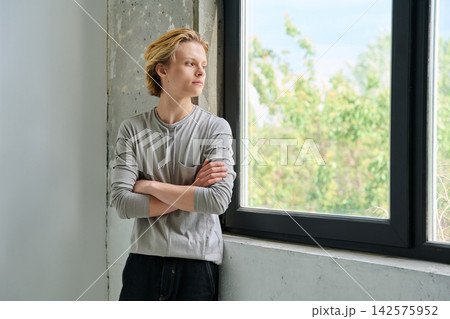 Thoughtful teenage boy with crossed arms by window Thoughtful teenage boy with crossed arms by window 142575952