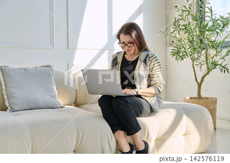 Portrait of middle aged woman using laptop sitting on sofa at home Portrait of middle aged woman using laptop sitting on sofa at home 142576119