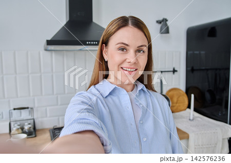 Cheerful woman in blue shirt taking selfie in bright modern kitchen 142576236