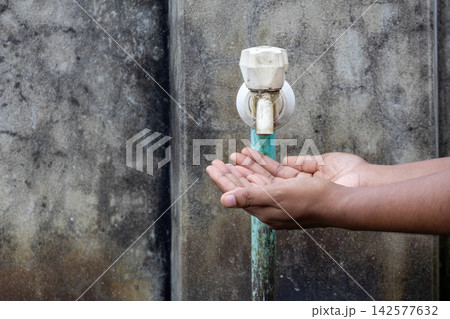 Human hands cupped under dry plastic water tap attached to weathered wall. Depicting water scarcity concept, global drought crisis, and resource shortage. Human hands cupped under dry plastic water tap attached to weathered wall. Depicting water scarcity concept, global drought crisis, and resource shortage. 142577632