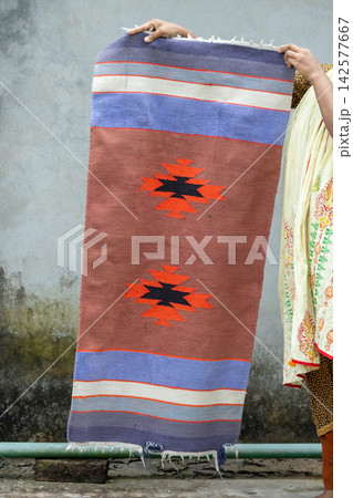 Handwoven traditional cotton satranji runner rug held by hands against rustic grey wall background. Ethnic geometric pattern floor mat for home decoration. 142577667