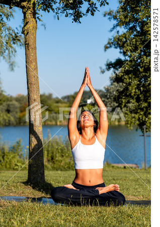 Hispanic Woman in Seated Backbend With Arms Overhead in a Summer Park. A young Hispanic woman practises a seated backbend with arms overhead by the tranquil lake in a summer park at dusk 142578571