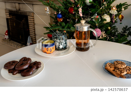 French Press Tea with Chocolate and Chip Cookies by Tree. A glass French press of tea with a candle and plates of chocolate gingerbread and chip cookies on a white table before a glowing Christmas tre French Press Tea with Chocolate and Chip Cookies by Tree. A glass French press of tea with a candle and plates of chocolate gingerbread and chip cookies on a white table before a glowing Christmas tre 142578637