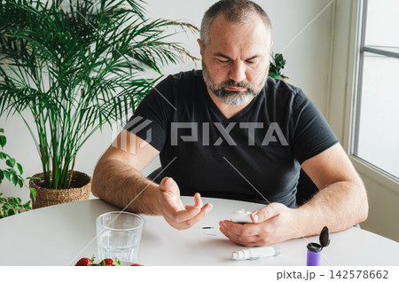 Mature Man Measuring Blood Sugar at Home with Fruit. Mature man measures his blood sugar with a glucometer at a table with a bowl of fresh strawberries, illustrating diabetes self-monitoring, a health 142578662