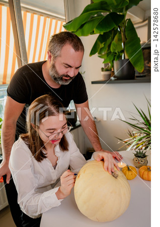 Father and Daughter Drawing on Pumpkin Together for Halloween at Home. Her father stands behind a daughter as she draws on a pale pumpkin at a festive table with mini pumpkins and a candle. A warm aut Father and Daughter Drawing on Pumpkin Together for Halloween at Home. Her father stands behind a daughter as she draws on a pale pumpkin at a festive table with mini pumpkins and a candle. A warm aut 142578680