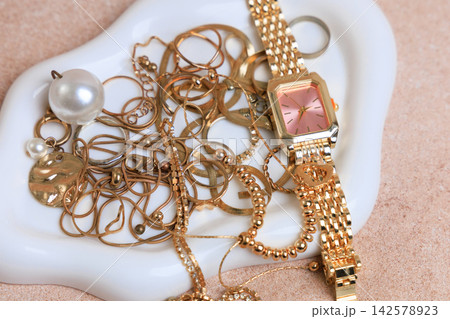 Various gold jewelry pieces and a watch arranged on a decorative plate in a well lit space during daytime 142578923