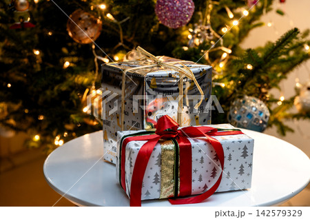 Stacked Christmas Gifts with Red Ribbon by Glowing Tree. Festive Christmas gifts wrapped in gold and white paper, one tied with a red ribbon and bow, stacked on a white table before a glowing Christma Stacked Christmas Gifts with Red Ribbon by Glowing Tree. Festive Christmas gifts wrapped in gold and white paper, one tied with a red ribbon and bow, stacked on a white table before a glowing Christma 142579329
