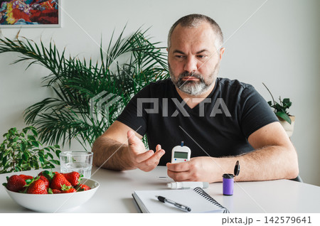Mature Man Doing a Finger Blood Sugar Test. Mature man does a finger- blood sugar test with a glucometer at a table with a bowl of fresh strawberries, illustrating diabetes self-monitoring and a healt Mature Man Doing a Finger Blood Sugar Test. Mature man does a finger- blood sugar test with a glucometer at a table with a bowl of fresh strawberries, illustrating diabetes self-monitoring and a healt 142579641