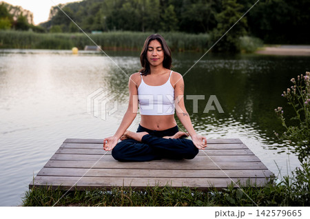 Young Woman in Seated Cross Legged Meditation in a Green Park. A young Hispanic woman practises a seated cross legged meditation in a green park beside a calm lake at sunset, nurturing wellness Young Woman in Seated Cross Legged Meditation in a Green Park. A young Hispanic woman practises a seated cross legged meditation in a green park beside a calm lake at sunset, nurturing wellness 142579665