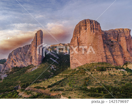 Rock Towers Rising Into a Smoke Filled Sky. Aerial view of the towering Mallos de Riglos rising into a sky filled with wildfire smoke in Aragon, Spain, the majestic red rock landmark of the Spanish Py Rock Towers Rising Into a Smoke Filled Sky. Aerial view of the towering Mallos de Riglos rising into a sky filled with wildfire smoke in Aragon, Spain, the majestic red rock landmark of the Spanish Py 142579697