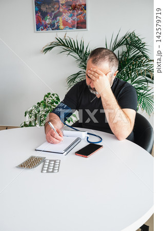 Worried Mature Man Measuring High Blood Pressure at Home. Worried mature man holds his head while measuring his blood pressure with a home monitor and writing in a notebook at a table with pills, illu Worried Mature Man Measuring High Blood Pressure at Home. Worried mature man holds his head while measuring his blood pressure with a home monitor and writing in a notebook at a table with pills, illu 142579719