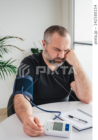 Man Measuring Blood Pressure with Home Arm Cuff Monitor. Mature man measures his blood pressure with a digital arm-cuff monitor at a table, illustrating hypertension self-monitoring, heart health Man Measuring Blood Pressure with Home Arm Cuff Monitor. Mature man measures his blood pressure with a digital arm-cuff monitor at a table, illustrating hypertension self-monitoring, heart health 142580154