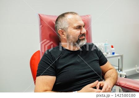 Mature Man Seated in Blood Donation Chair at Clinic. Mature man relaxing in a red blood donation chair at a clinic, looking aside in a calm moment before giving blood, illustrating voluntary blood Mature Man Seated in Blood Donation Chair at Clinic. Mature man relaxing in a red blood donation chair at a clinic, looking aside in a calm moment before giving blood, illustrating voluntary blood 142580180