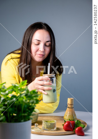 Smiling Woman Sipping Matcha Latte with Straw, Summer. Cheerful woman sipping green matcha latte through straw with content smile in cozy summer setting. Rustic wooden background with ceramic bowl 142580237
