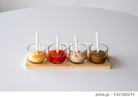 Mustard and dipping sauces row on bamboo board side view. Side view of mustard and dipping sauces arranged in row on bamboo board with white spoons, gourmet condiment lineup for BBQ grilling and food Mustard and dipping sauces row on bamboo board side view. Side view of mustard and dipping sauces arranged in row on bamboo board with white spoons, gourmet condiment lineup for BBQ grilling and food 142580243