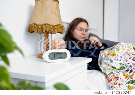Alarm Clock on Nightstand in Teen Bedroom, Morning School Routine. Close-up of alarm clock and smartphone on bedside table in teenager bedroom. Soft morning light, glasses and lamp in blurred backgrou Alarm Clock on Nightstand in Teen Bedroom, Morning School Routine. Close-up of alarm clock and smartphone on bedside table in teenager bedroom. Soft morning light, glasses and lamp in blurred backgrou 142580244