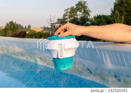 Person is using a floating chlorine dispenser with an integrated thermometer to check and maintain the hygiene and temperature of an above ground swimming pool during summer season Person is using a floating chlorine dispenser with an integrated thermometer to check and maintain the hygiene and temperature of an above ground swimming pool during summer season 142580361